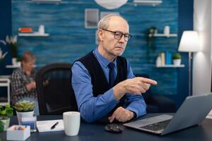 Thoughtfull senior businessman pointing at laptop whille working from home. Elderly man entrepreneur in home workplace using portable computer sitting at desk while wife is holding tv remote. photo