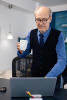 Caucasian old man enjoying a cup of coffee using laptop wearing glasses.Elderly man entrepreneur in home workplace using portable computer sitting at desk while wife is holding tv remote. photo