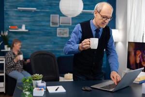 Retired businessman turning on laptop enjoying a cup of coffee. Elderly man entrepreneur in home workplace using portable computer sitting at desk while wife is holding tv remote. photo