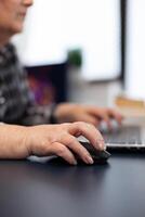 Close up of old entrepreneur lady using mouse in home office. Elderly woman in home living room using moder technoloy laptop for communication sitting at desk indoors. photo