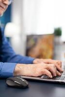 Close up of senior man hands typing on laptop keyboard. Elderly man entrepreneur in home workplace using portable computer sitting at desk while wife is holding tv remote photo
