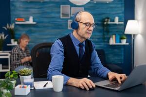 Senior businessman listening music wearing headphones working on laptop. Elderly man entrepreneur in home workplace using portable computer sitting at desk while wife is reading a book sitting on sofa. photo