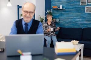 Old woman relaxing on couch holding a cup of coffee and husband is working. Elderly man entrepreneur in home workplace using portable computer sitting at desk while wife is holding tv remote. photo