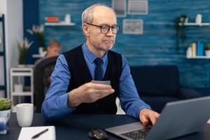 Senior man using credit card to check bank account. Elderly man checking online banking to make shppping payment looking at laptop while wife is reading a book sitting on sofa. photo