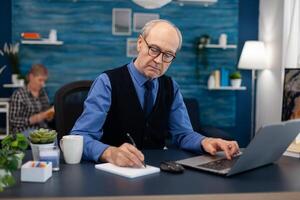 Senior businessman writing notes wearing glasses working in home office. Elderly man entrepreneur in home workplace using portable computer sitting at desk while wife is reading a book sitting on sofa. photo