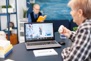 Senior pensioner listening doctor talking about treatment during online consultation. Elderly woman discussing with healthcare practitioner in the course of remote call and husband is reading a book on sofa. photo