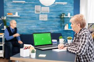 Senior woman taking notes on notebook looking at portable computer with copy space available. Elderly woman working on laptop with green screen and husband holding tv remote control. photo