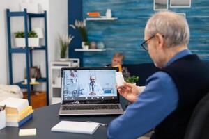Senior man showing doctor pills bottle at webcam during call. Elderly man discussing with healthcare practitioner in the course of remote call and wife is reading a book on sofa. photo