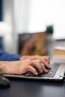 Close up of senior man hands typing on laptop keyboard. Elderly man entrepreneur in home workplace using portable computer sitting at desk while wife is holding tv remote photo