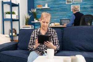 Middle aged lady relaxing on sofa reading on tablet pc anjoying content. Elderly woman using moder technoloy tablet pc in home living room and husband working on laptop computer photo
