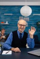 Smart senior businessman working on laptop wearing a tie and glasses. Elderly man entrepreneur in home workplace using portable computer sitting at desk while wife is holding tv remote. photo