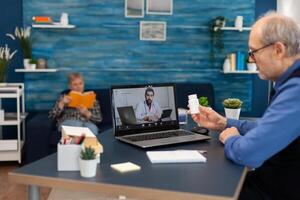 Young doctor examining senior man during conference. Elderly man discussing with healthcare practitioner in the course of remote call and wife is reading a book on sofa. photo