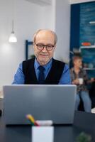 Old woman relaxing on couch holding a cup of coffee and husband is working. Elderly man entrepreneur in home workplace using portable computer sitting at desk while wife is holding tv remote. photo