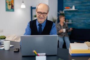 Old woman relaxing on couch holding a cup of coffee and husband is working. Elderly man entrepreneur in home workplace using portable computer sitting at desk while wife is holding tv remote. photo