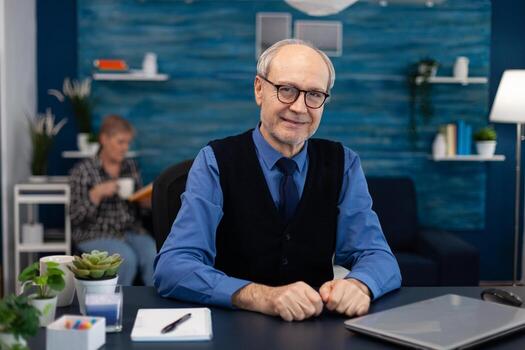 Portrait of senior man sitting at desk office looking at camera. Elderly man entrepreneur in home workplace using portable computer sitting at desk while wife is reading a book sitting on sofa. photo