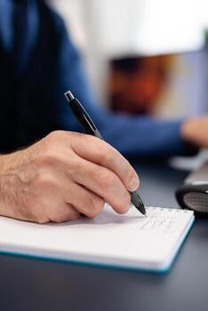 Close up of man taking notes on notebook while working from home. Elderly man entrepreneur in home workplace using portable computer sitting at desk while wife is reading a book sitting on sofa. photo