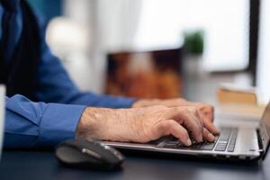 Close up of senior man hands typing on laptop keyboard. Elderly man entrepreneur in home workplace using portable computer sitting at desk while wife is holding tv remote photo
