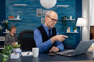 Thoughtfull senior businessman pointing at laptop whille working from home. Elderly man entrepreneur in home workplace using portable computer sitting at desk while wife is holding tv remote. photo