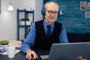 Senior businessman listening music wearing headphones working on laptop. Elderly man entrepreneur in home workplace using portable computer sitting at desk while wife is reading a book sitting on sofa. photo