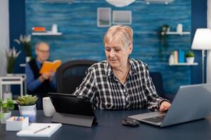 Senior business owner working from home office using portable computer. Elderly woman in home living room using moder technoloy laptop tablet pc for communication sitting at desk indoors. photo