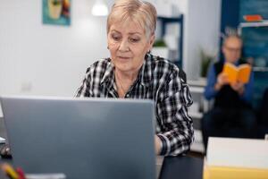 Senior lady entrepreneur working and pointing at computer screen while browsing on internet. Elderly woman in home living room using moder technoloy laptop for communication sitting at desk indoors. photo