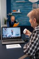 Senior pensioner listening doctor talking about treatment during online consultation. Elderly woman discussing with healthcare practitioner in the course of remote call and husband is reading a book on sofa. photo