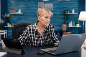 Senior business owner working from home office using portable computer. Elderly woman in home living room using moder technoloy laptop tablet pc for communication sitting at desk indoors. photo