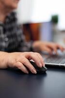 Close up of old entrepreneur lady using mouse in home office. Elderly woman in home living room using moder technoloy laptop for communication sitting at desk indoors. photo