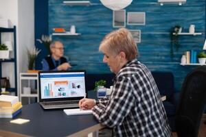 Elderly senior woman taking notes in notebook while working on laptop in cozy living room. Adult internet finance computer caucasian mature technology glasses browsing connection retored retirement photo