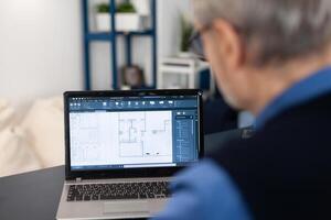 Overshoulder shot of senior architect working on laptop to finish a project. Experienced construction engineer designer looking at house blueprints on laptop computer from home office. photo