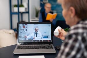 Older woman listening doctor indication about health treatment during call. Elderly woman discussing with healthcare practitioner in the course of remote call and husband is reading a book on sofa. photo