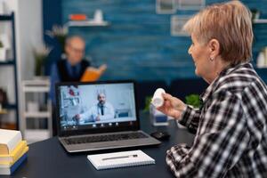 Senior woman showing pills bottle during medical online conference. Elderly woman discussing with healthcare practitioner in the course of remote call and husband is reading a book on sofa. photo