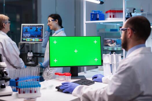 Biochemist working in laboratory using green mock-up screen for biochemistry experiment with chroma key monitor. Team of microbiologists doing vaccine analysis and discovery treatment cure against covid19 photo