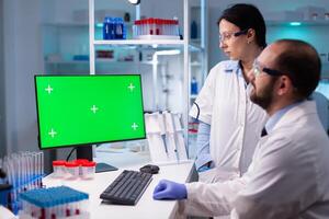 Modern medical research laboratory with two scientists using computer with green chroma key screen. Doctors specialists discussing innovative treatment against covid19 using high technology equipment. photo