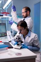 Scientist examining blood sample looking under microscope while chemist doctor using micropipette for pharmaceutical expertise. Team of medical researchers working to discovering cure against disease photo