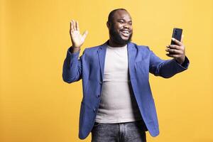 Cheerful man excited to greet friends during teleconference meeting using smartphone, studio background. Smiling person waving hand, saluting mate during internet call on mobile phone photo