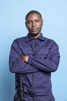Portrait of serious proud security guard posing in uniform on camera, standing with arms crossed against blue background. African american armed bodyguard works on ensuring protection. photo