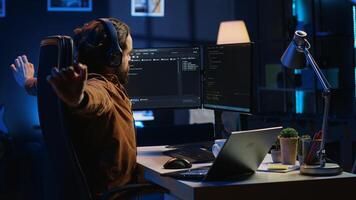 Software programmer taking break from coding, putting headphones on and stretching on desk chair during remote job shift. IT professional pausing tasks, listening music, loosening up, camera A photo