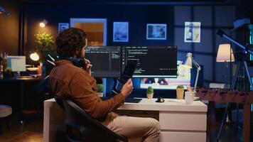 Software developer programming in home office, crosschecking lines of code between tablet and PC screen. IT admin holding portable device, using it as reference while coding on computer, camera B photo