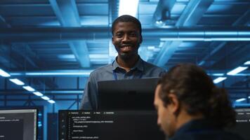 Portrait of cheerful african american manager supervising team in server room providing computing resources for workloads. Jolly supervisor oversees technicians in data center mending supercomputers photo