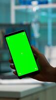 Close up of biochemist holding and looking at phone with chroma key display standing in biological lab while nurse in white coat bringing blood sample. Scientist using phone with mockup, green screen photo