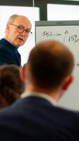 Elderly businessman giving presentation to multiethnic team of colleagues using flip chart, drawing, explaning financial graphs in meeting room. Manager interacting with audience at corporate workshop photo