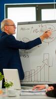 Senior businessman in eyeglasses and formal suit drawing financial plan on flipchart and explaining to interracial team of colleagues while giving presentation during conference meeting, brainstorming photo