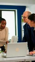 Young multicultural workers group talking, looking at laptop and analyzing digital datas sitting at workplace desk. Professional colleagues team discussing project using devices in corporate office. photo