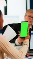 Back view of black businesswoman sitting at conference desk holding smartphone with green screen while colleagues working on background. Multiethnic coworkers planning project on chroma key display photo