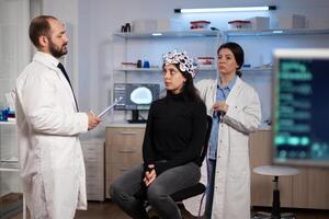 Scientist doctor discussing neurology disease symptoms with woman patient while neurologist researcher adjusting eeg scanner during neuroscience experiment in lab. Specialist analyzing brain activity photo
