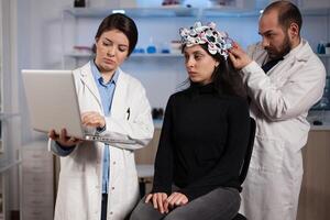 Specialist doctor holding laptop computer showing tomography to woman patient while neurologist man adjusting eeg scanner during neurology experiment. Team of researchers analyzing brain activity photo