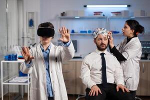 Neurologist doctor wearing virtual reality headset while researcher woman adjusting eeg scanner of man patient analyzing brain activity during neurology experiment. Scientist engineer using high tech photo