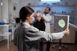 Neurologist woman doctor looking at monitor with medical tomography analyzing brain activity using high tech equipment. In background specialist man adjusting eeg headset of woman patient photo