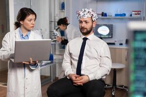 Researcher woman doctor typing neurological disease symptoms on laptop discussing medical treatment with patient. Neurologist engineer monitoring nervous system activity of man with eeg headset photo